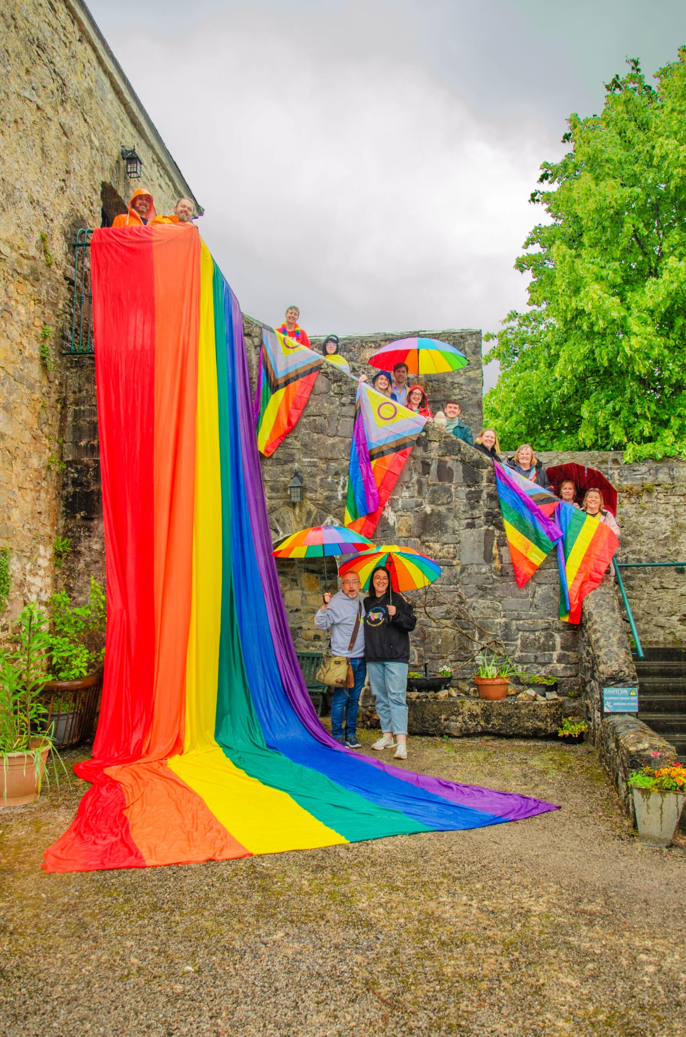 A group of Roscommon Pride volunteers with colourful Pride flags and umbrellas. A large rainbow flag cascades down the building.