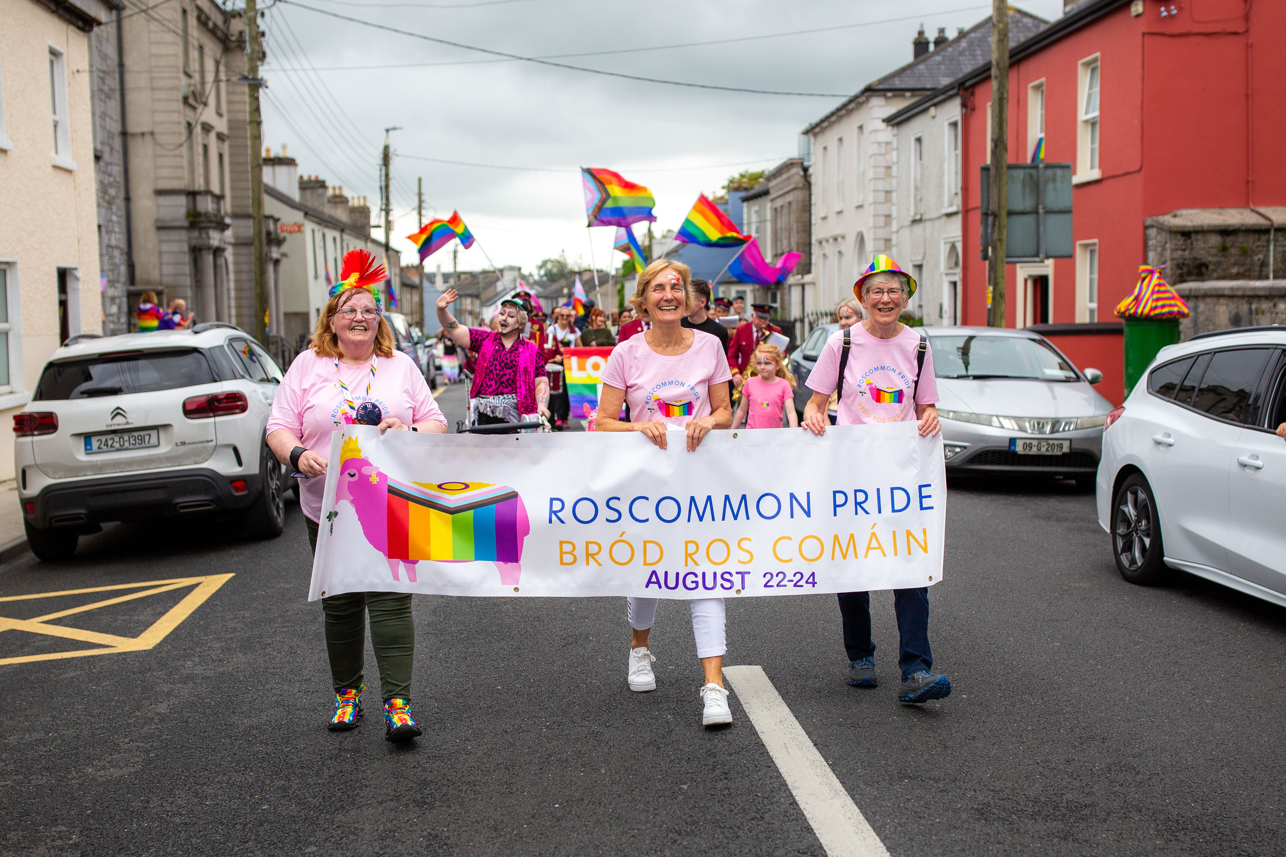 Participants celebrating Pride, dressed in pink t-shirts and rainbow accessories, march down the street holding a banner that reads "Roscommon Pride, Bród Ros Comáin, August 22-24".