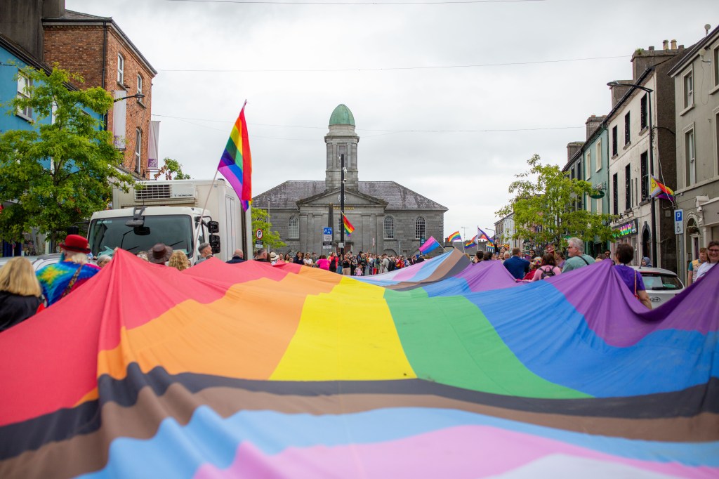 A vibrant crowd holds a large rainbow flag during a Pride march, with Roscommon's Harrison Hall in the background and rainbow flags displayed along the streets.