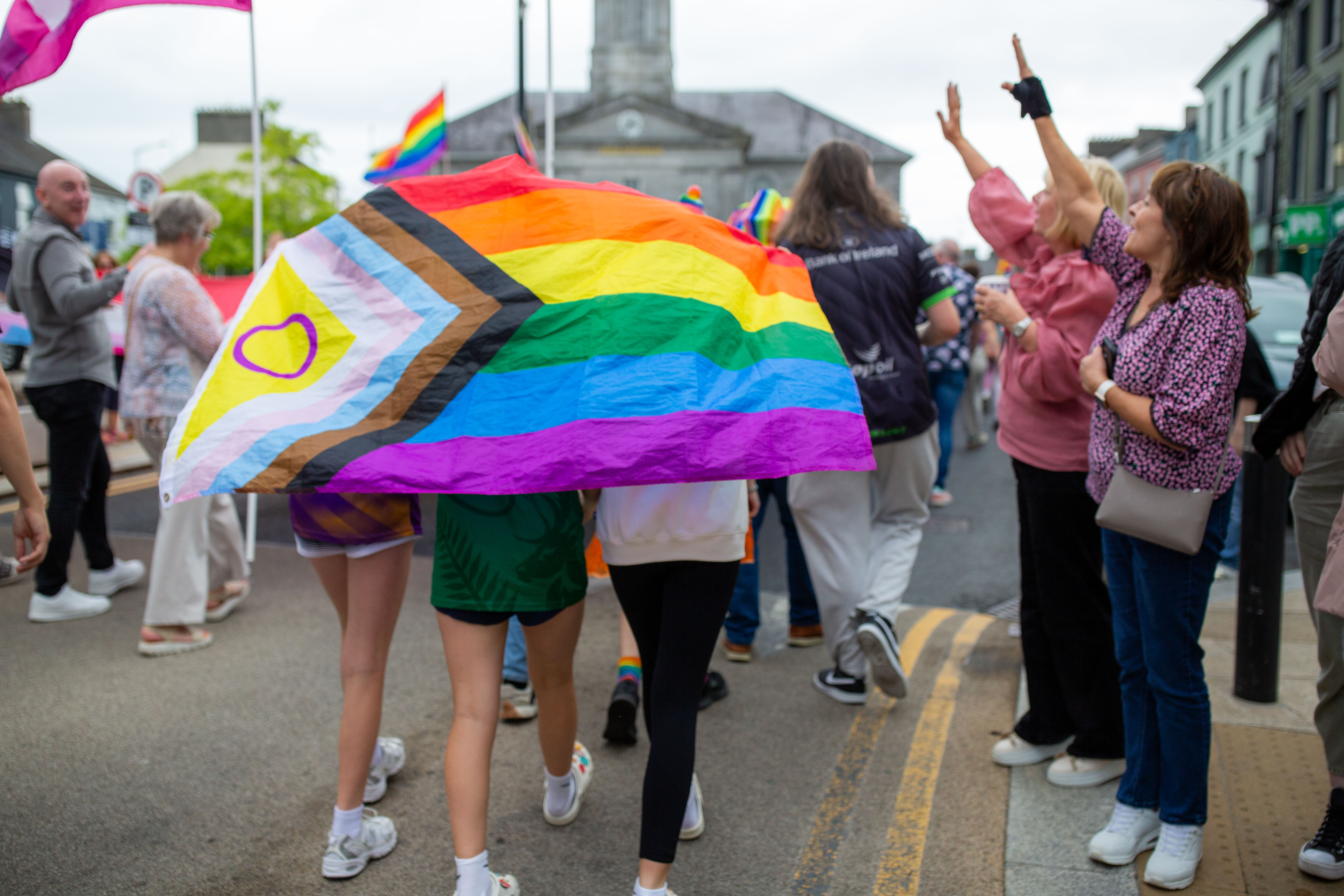 A group of people walking in a Pride march, with one person holding a large rainbow flag and others in the background waving and showing support.